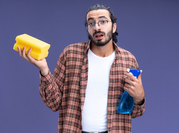 Onder de indruk jonge knappe schoonmaakster die de spuitfles van de t-shirtholding met spons draagt die op blauwe muur wordt geïsoleerd