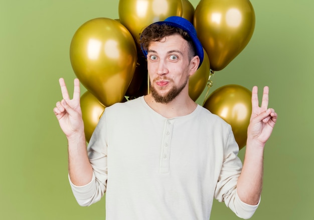 Onder de indruk jonge knappe Slavische feestjongen die feestmuts draagt die zich achter ballonnen bevinden die camera bekijken die vredestekens doen die op olijfgroene achtergrond worden geïsoleerd