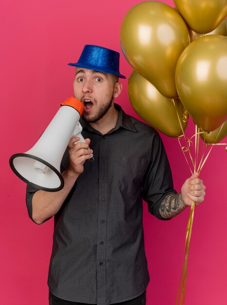 Onder de indruk jonge knappe Slavische feestmens die feestmuts draagt die ballonnen houdt die spreker in de buurt van mond houdt die camera bekijkt die op karmozijnrode achtergrond wordt geïsoleerd