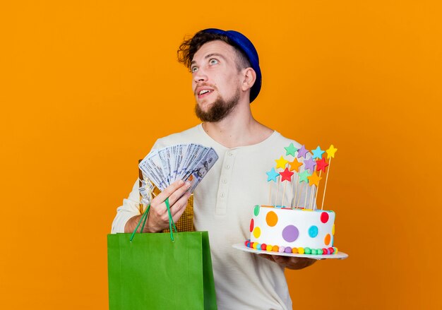 Onder de indruk jonge knappe Slavische partij kerel met feestmuts bedrijf geschenkdoos geld papieren zak en verjaardagstaart met sterren opzoeken geïsoleerd op een oranje achtergrond met kopie ruimte