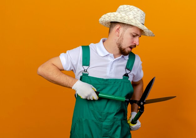 Onder de indruk jonge knappe Slavische tuinman in uniform met hoed en tuinhandschoenen houden en kijken naar geïsoleerde snoeischaren