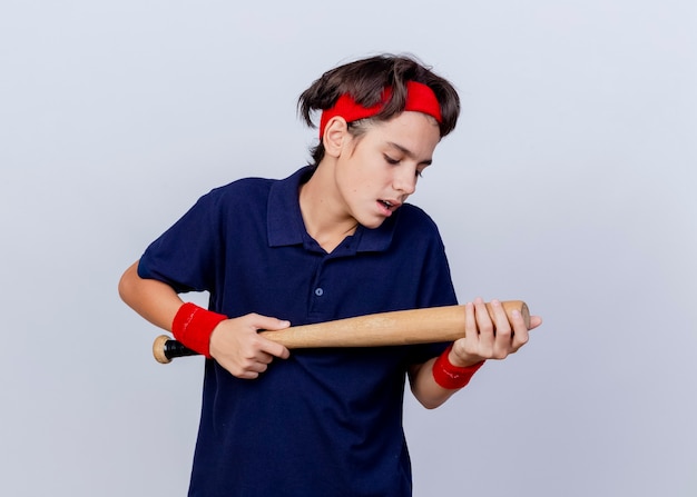 Onder de indruk jonge knappe sportieve jongen die hoofdband en polsbandjes met beugels draagt die en honkbalknuppel houden die op witte muur wordt geïsoleerd