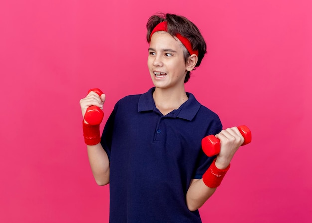Onder de indruk jonge knappe sportieve jongen die hoofdband en polsbandjes met beugels draagt die halters houden die naar kant kijken die op roze muur met exemplaarruimte wordt geïsoleerd
