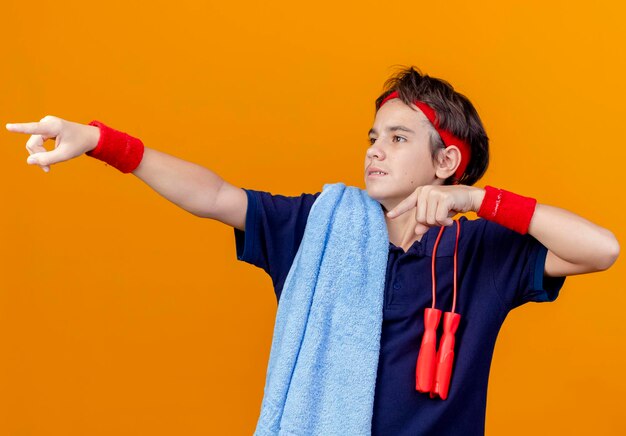 Onder de indruk jonge knappe sportieve jongen met hoofdband en polsbandjes met beugels en handdoek met springtouw op schouders kijken en wijzend naar kant geïsoleerd op oranje muur