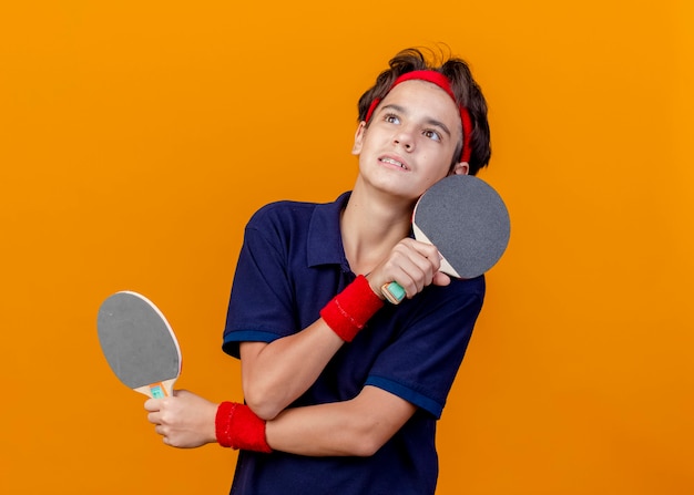 Onder de indruk jonge knappe sportieve jongen met hoofdband en polsbandjes met beugels pingpong rackets houden handen gekruist opzoeken geïsoleerd op oranje muur