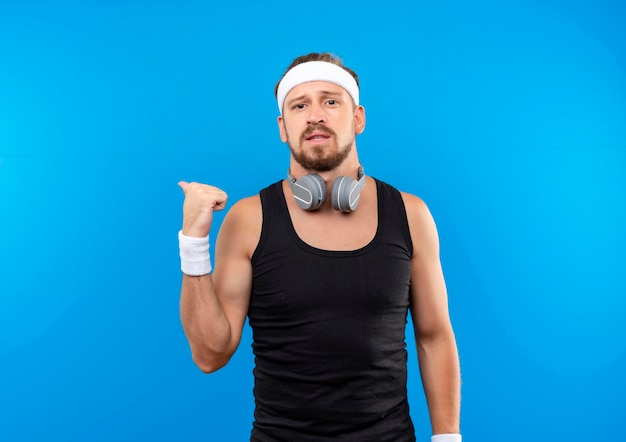 Onder de indruk jonge knappe sportieve man met hoofdband en polsbandjes en koptelefoon om de nek wijzend achter geïsoleerd op blauwe muur