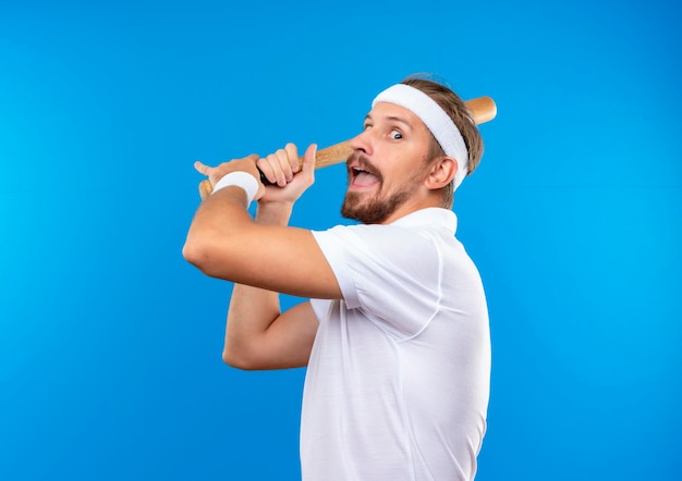 Onder de indruk jonge knappe sportieve man met hoofdband en polsbandjes met honkbalknuppel en klaar om de bal te raken geïsoleerd op blauwe muur