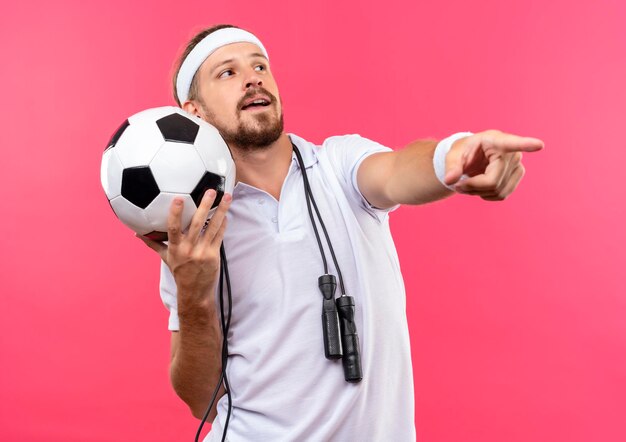 Onder de indruk jonge knappe sportieve man met hoofdband en polsbandjes met voetbal kijkend naar de zijkant en recht wijzend met springtouw om nek geïsoleerd op roze muur