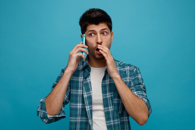 Onder de indruk jonge man praten aan de telefoon kijkend naar de zijkant houden hand in de buurt van mond fluisteren geïsoleerd op blauwe achtergrond