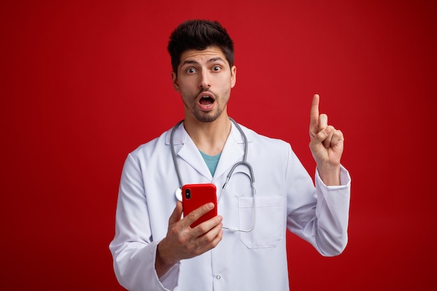 Onder de indruk jonge mannelijke arts dragen medisch uniform en stethoscoop om zijn nek met mobiele telefoon kijken camera omhoog geïsoleerd op rode achtergrond