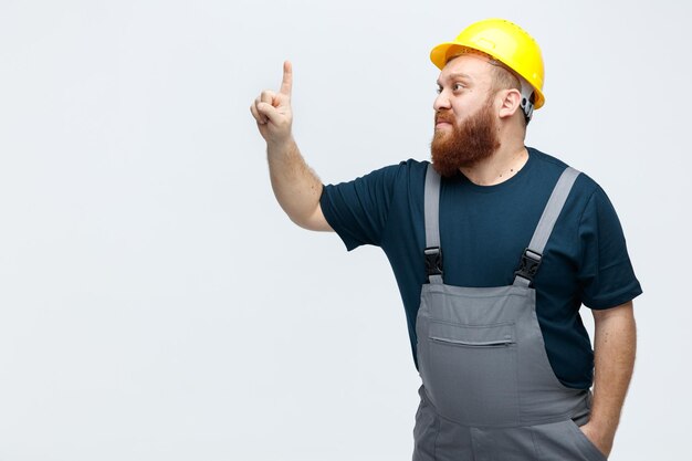 Onder de indruk jonge mannelijke bouwvakker met veiligheidshelm en uniform kijkend naar de zijkant die omhoog wijst terwijl hij een andere hand in de zak houdt geïsoleerd op een witte achtergrond met kopieerruimte