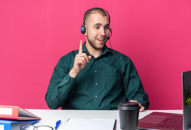 Onder de indruk jonge mannelijke callcenter-operator met een headset die aan het bureau zit met kantoorhulpmiddelen die naar laptoppunten kijken naar boven