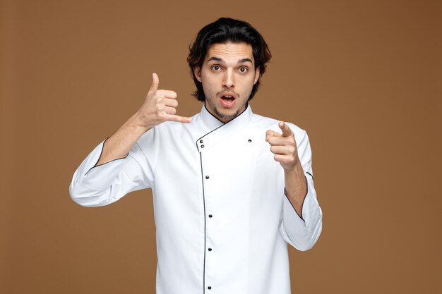 onder de indruk jonge mannelijke chef-kok die een oproepgebaar draagt, kijkend en wijzend op camera-uniform geïsoleerd op bruine achtergrond