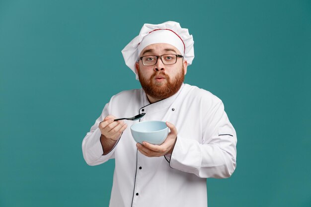 Onder de indruk jonge mannelijke chef-kok met een uniforme bril en pet met lege kom en lepel kijkend naar camera geïsoleerd op blauwe achtergrond