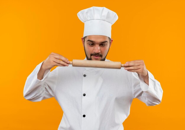 Onder de indruk jonge mannelijke kok in chef-kok uniform houden en kijken naar deegroller geïsoleerd op oranje muur