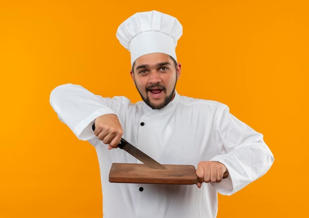 Onder de indruk jonge mannelijke kok in uniform van de chef-kok met mes en snijplank geïsoleerd op oranje muur