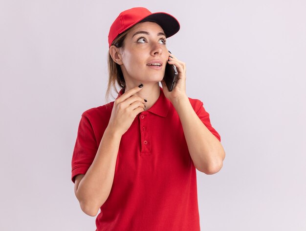 Onder de indruk jonge mooie bezorger in uniforme gesprekken aan de telefoon en kijkt naar kant geïsoleerd op een witte muur