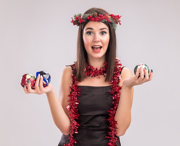 Onder de indruk jonge mooie blanke meisje dragen kerst hoofd krans en klatergoud slinger rond nek houden kerstballen kijken camera geïsoleerd op witte achtergrond