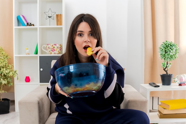 Onder de indruk jonge mooie blanke vrouw zittend op een fauteuil in de ontworpen woonkamer met kom met chips op zoek en het eten van aardappelchips