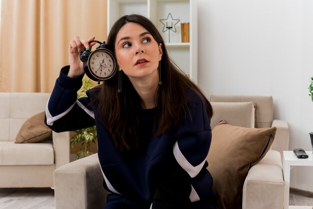 Onder de indruk jonge mooie blanke vrouw zittend op een fauteuil in de ontworpen woonkamer met wekker in de buurt van oor kijken naar kant