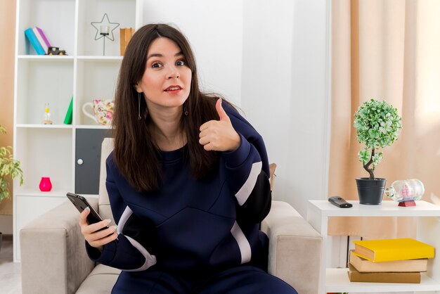 Onder de indruk jonge mooie blanke vrouw zittend op een fauteuil in ontworpen woonkamer met mobiele telefoon op zoek en duim opdagen