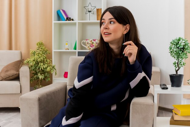 Onder de indruk jonge mooie blanke vrouw zittend op een stoel in ontworpen woonkamer elleboog op fauteuil houden hand in de lucht kijken kant