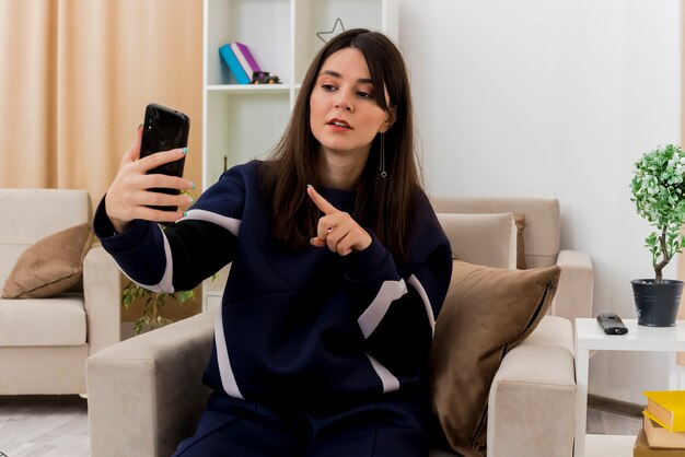 Onder de indruk jonge mooie blanke vrouw zittend op een stoel in ontworpen woonkamer houden en kijken naar mobiele telefoon en erop wijzen
