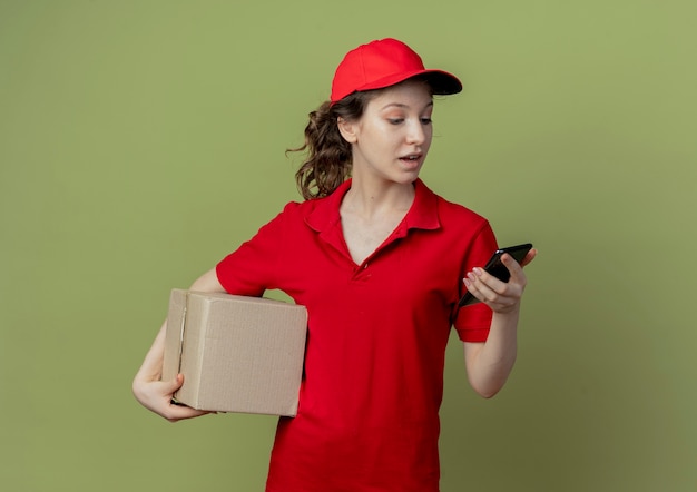 Onder de indruk jonge mooie levering meisje in rood uniform en pet bedrijf kartonnen doos en mobiele telefoon kijken naar telefoon geïsoleerd op olijfgroene achtergrond met kopie ruimte