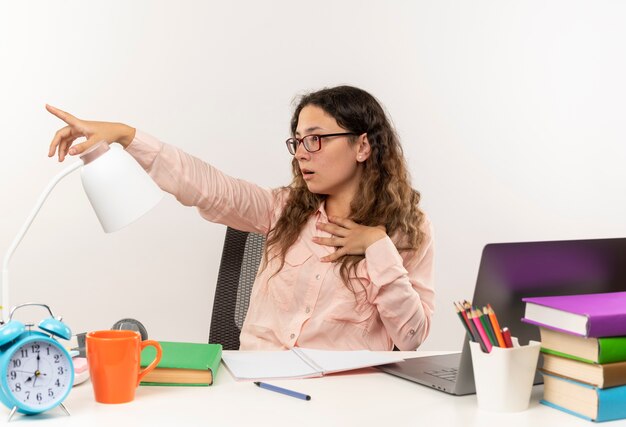 Onder de indruk jonge mooie schoolmeisje bril zit aan Bureau met school tools haar huiswerk kijken en wijzend naar kant met hand op borst geïsoleerd op wit