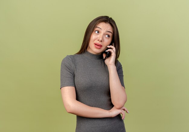 Onder de indruk jonge mooie vrouw kant kijken en praten over telefoon geïsoleerd op olijfgroene achtergrond met kopie ruimte