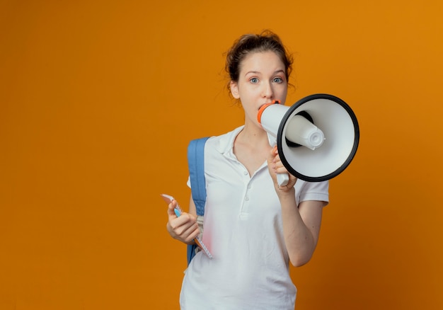 Gratis foto onder de indruk jonge mooie vrouwelijke student die rugtas draagt die pen en notitieblok houdt die door spreker spreken die op oranje achtergrond met exemplaarruimte wordt geïsoleerd