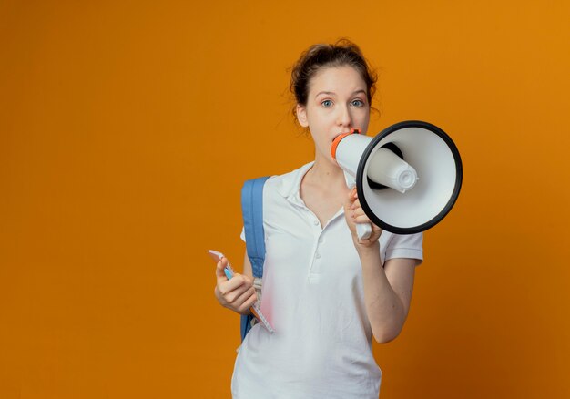 Onder de indruk jonge mooie vrouwelijke student die rugtas draagt die pen en notitieblok houdt die door spreker spreken die op oranje achtergrond met exemplaarruimte wordt geïsoleerd