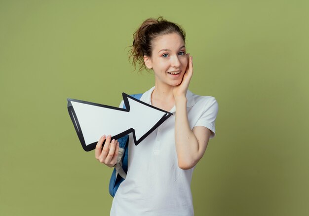 Onder de indruk jonge mooie vrouwelijke student draagt ?? rugtas met pijlteken die naar de zijkant wijst en het aanraken van gezicht geïsoleerd op olijfgroene achtergrond met kopie ruimte