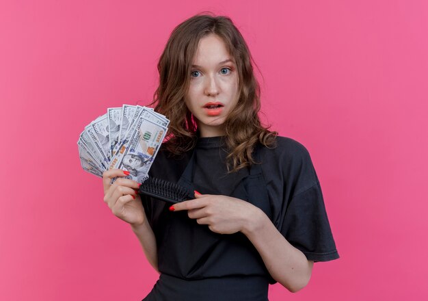 Onder de indruk jonge Slavische vrouwelijke kapper die eenvormige holdingskam en geld draagt die op roze achtergrond wordt geïsoleerd