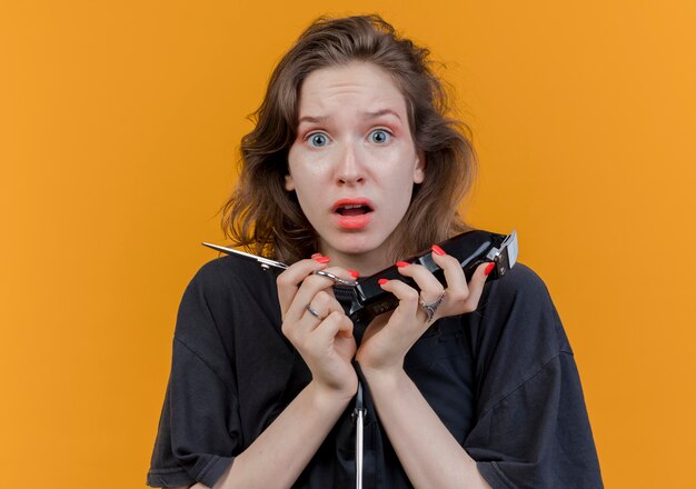 Onder de indruk jonge Slavische vrouwelijke kapper die eenvormige holdingsschaar en tondeuses dragen die camera bekijken die op oranje achtergrond wordt geïsoleerd