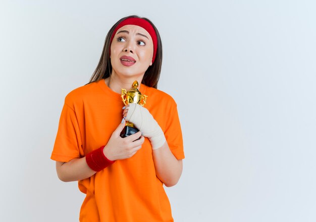 Onder de indruk jonge sportieve vrouw die hoofdband en polsbandjes draagt die de winnaarkop van de zijholding bekijken die op witte muur met exemplaarruimte wordt geïsoleerd