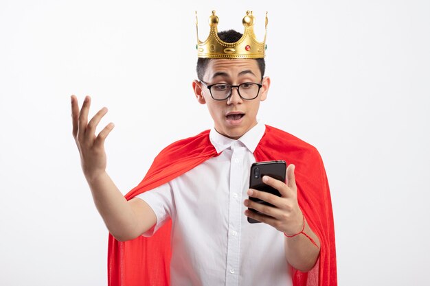 Onder de indruk jonge superheld jongen in rode cape bril en kroon houden en kijken naar mobiele telefoon houden hand in lucht geïsoleerd op witte achtergrond