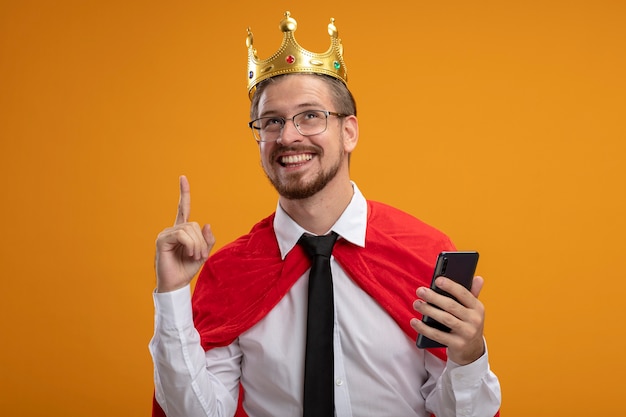 Gratis foto onder de indruk jonge superheld man met stropdas en kroon met bril met telefoon en wijst naar omhoog geïsoleerd op een oranje achtergrond