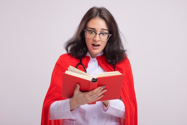 Onder de indruk jonge superheld vrouw in rode cape dragen dokter uniform en stethoscoop houden en lezen boek geïsoleerd op een witte muur met kopie ruimte