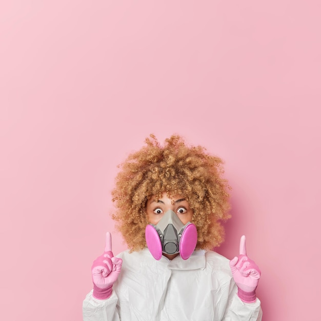 Onder de indruk jonge vrouw met krullend haar in gasmasker beschermend pak waarschuwt voor gevaar vertelt over wereldwijde catastrofepunten hierboven op lege ruimte geïsoleerd over roze achtergrond Coronavirus-infectie