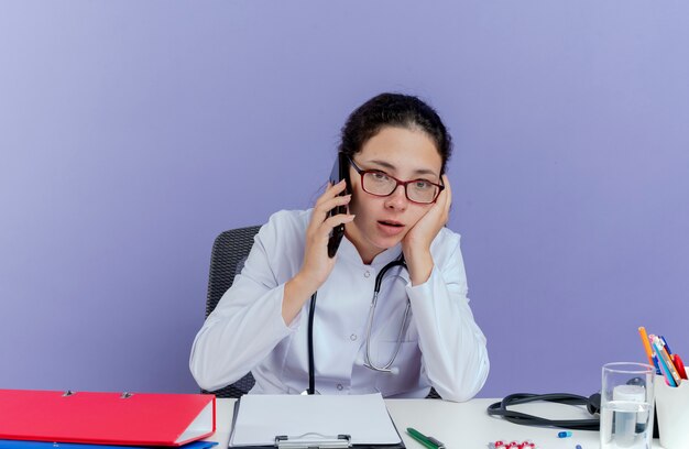 Onder de indruk jonge vrouwelijke arts die medische mantel en stethoscoop draagt die aan bureau zit met medische hulpmiddelen die op telefoon spreken die rechtstreeks aanraken gezicht geïsoleerd kijkt