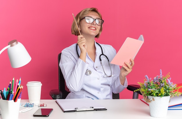 Onder de indruk jonge vrouwelijke arts met medische mantel met stethoscoop en bril zit aan bureau met medische hulpmiddelen met notitieboekje met potlood geïsoleerd op roze muur