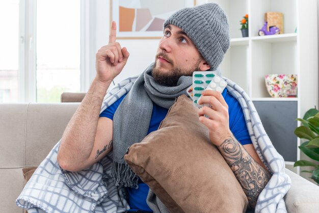 Onder de indruk jonge zieke man met sjaal en wintermuts gewikkeld in deken zittend op de bank in de woonkamer met kussen en pakjes pillen kijkend en omhoog wijzend