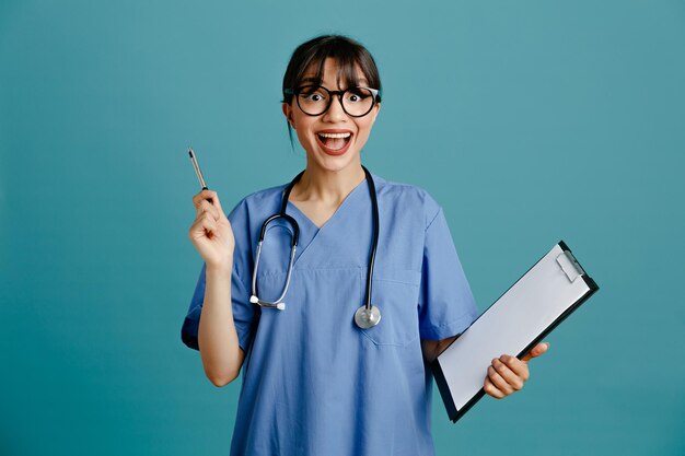 Onder de indruk klembord met pen jonge vrouwelijke arts dragen van uniforme fith stethoscoop geïsoleerd op blauwe achtergrond