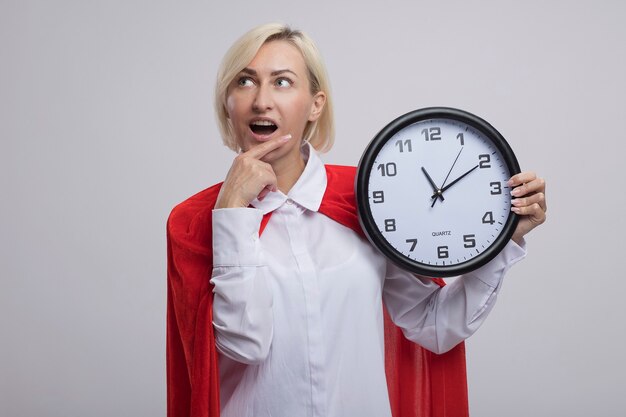 Onder de indruk middelbare leeftijd blonde superheld vrouw in rode cape met klok hand zetten kin kijken naar kant geïsoleerd op een witte muur met kopie ruimte