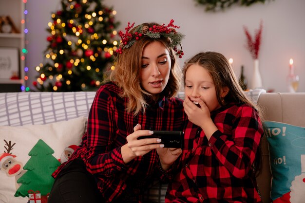 Onder de indruk moeder en dochter kijken naar iets op telefoon zittend op de bank en genieten van kersttijd thuis