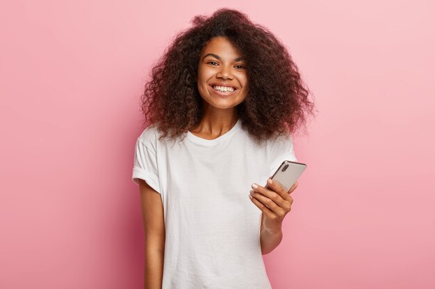 Onder de indruk mooie Afro-vrouw met luxe krullend haar, houdt moderne mobiele telefoon vast, wacht op oproep