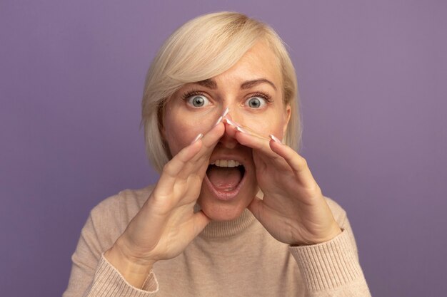 Onder de indruk mooie blonde Slavische vrouw houdt handen dicht bij mond geïsoleerd op paarse muur