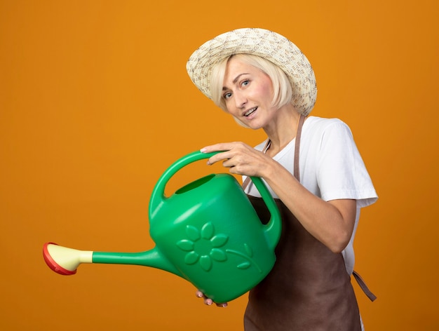 Onder de indruk tuinman vrouw van middelbare leeftijd in tuinman uniform staande in profiel weergave dragen hoed bedrijf gieter doen alsof drenken bloemen kijken naar voorzijde geïsoleerd op oranje muur