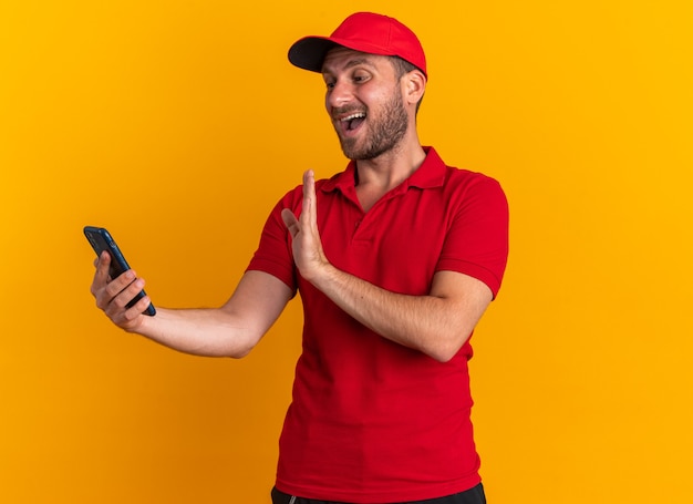 Onder de indruk van de jonge blanke bezorger in rood uniform en pet die mobiele telefoon vasthoudt en kijkt terwijl hij hallo gebaar doet geïsoleerd op een oranje muur